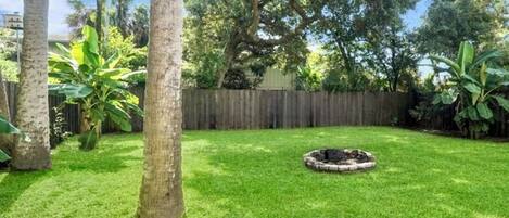 A backyard with a well-maintained lawn, a circular fire pit, and tall trees.