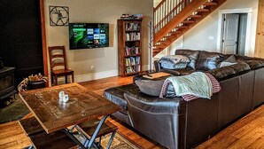 Una sala de estar acogedora con un sofá de cuero, una mesa de centro de madera y una estantería. Un televisor está montado en la pared, y se ve una escalera al fondo.