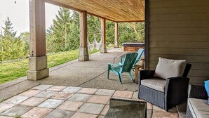 Un patio al aire libre con techo de madera, pilares de concreto y vista a un jardín. Hay dos sillas y una mesa.
