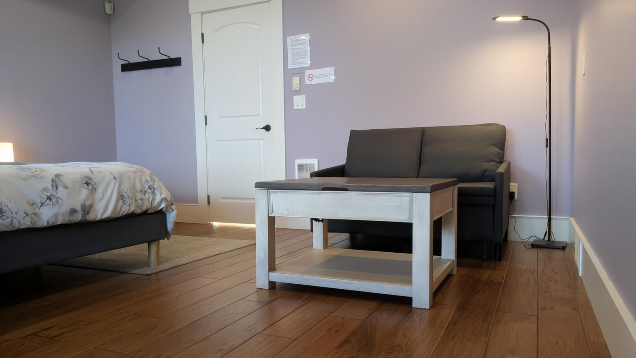 A bedroom with a bed, a small white table, a black chair, and a floor lamp.