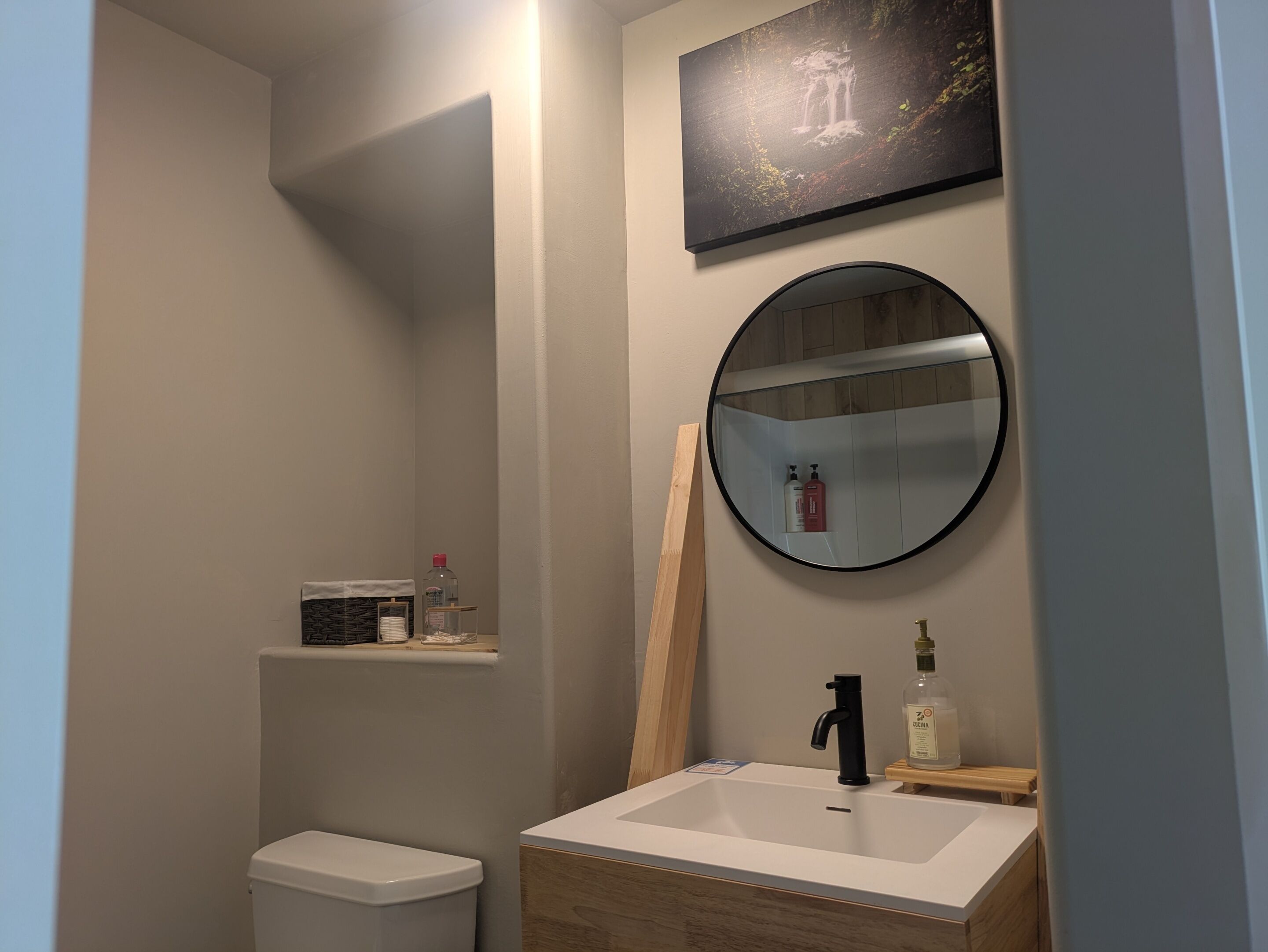 Un baño con inodoro, lavamanos con espejo y repisas con artículos de aseo.