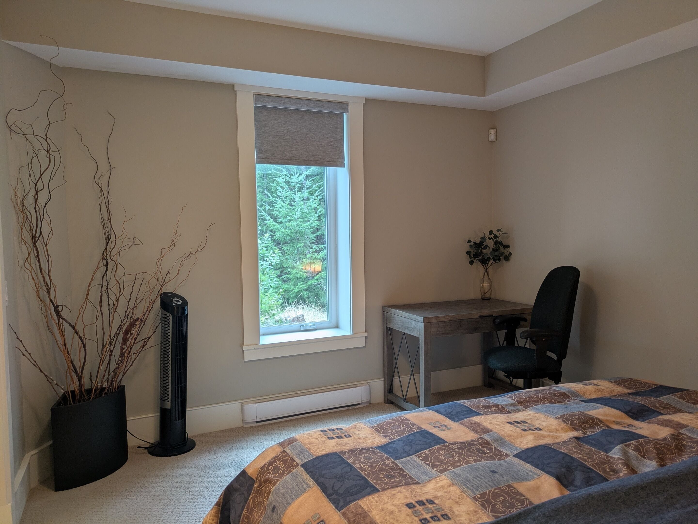 Un dormitorio con cama, escritorio, silla, radiador y ventana con vista a árboles.