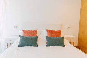 A neatly made bed with white bedding and two pillows, one orange and one green, in a bright room with white walls and a wooden door.