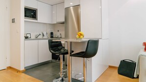 A modern kitchen with white cabinets, a stainless steel refrigerator, a microwave, and a fruit bowl on the counter. There are two black bar stools and a green bag on the floor. - Oporto Yellow Apartments104 (Porto)