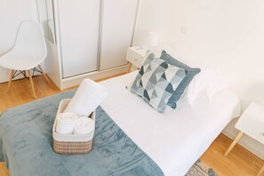 Una cama bien hecha con un edredón azul, una almohada con diseño geométrico y una mesita de noche con una lámpara. Se observa una silla blanca y una pequeña mesa con toallas y una cesta.