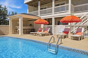 A swimming pool with lounge chairs and umbrellas, a covered seating area, and a multi-story building with balconies.