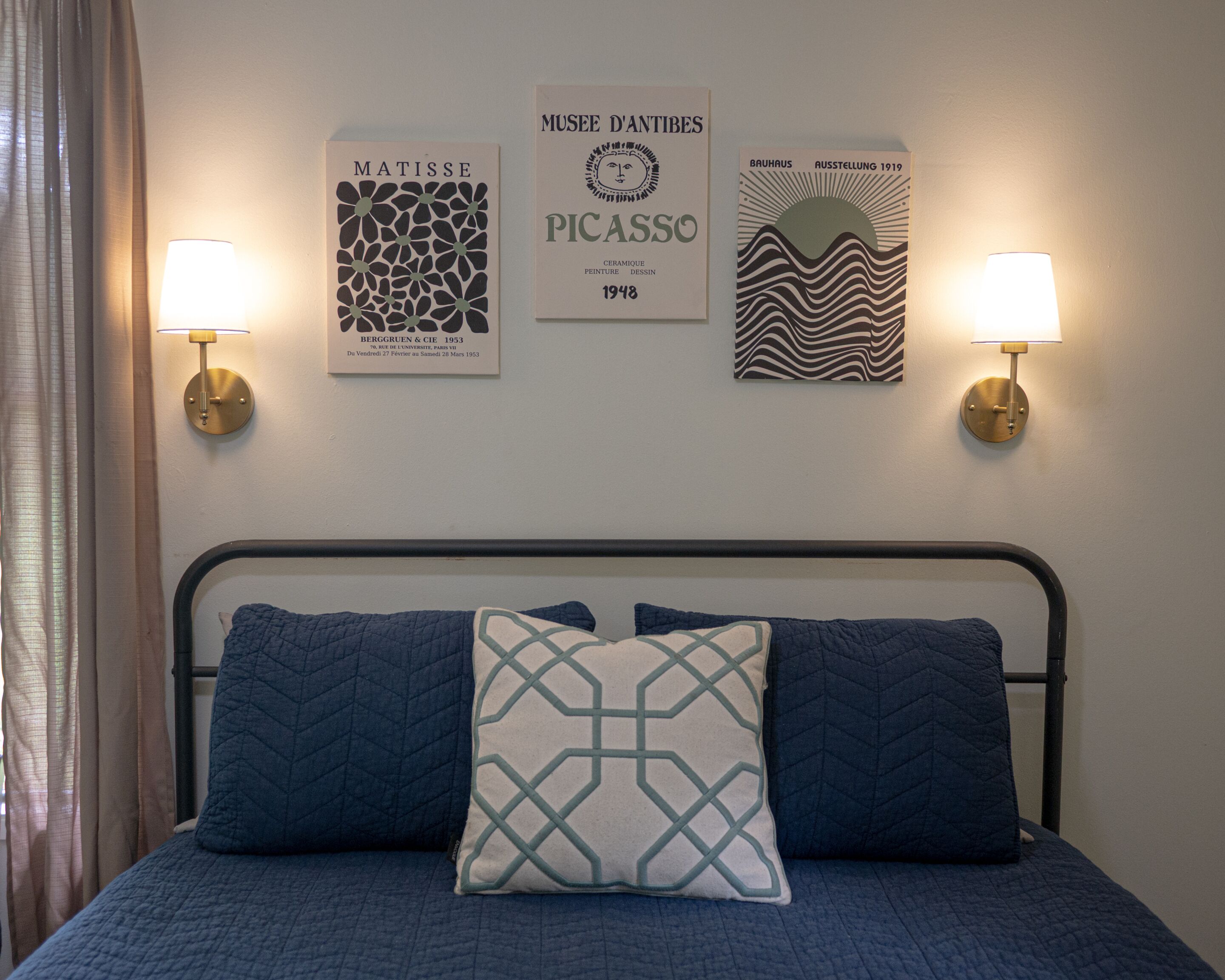 A bed with a metal frame and blue bedding, two wall-mounted lamps, and three framed artworks on the wall.