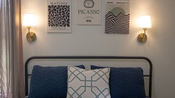 A bed with a metal frame and blue bedding, two wall-mounted lamps, and three framed artworks on the wall.