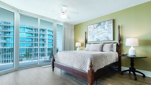 Une chambre d’hôtel avec un grand lit, deux tables de chevet, un ventilateur de plafond et une vue sur un immeuble à travers de grandes fenêtres du sol au plafond.