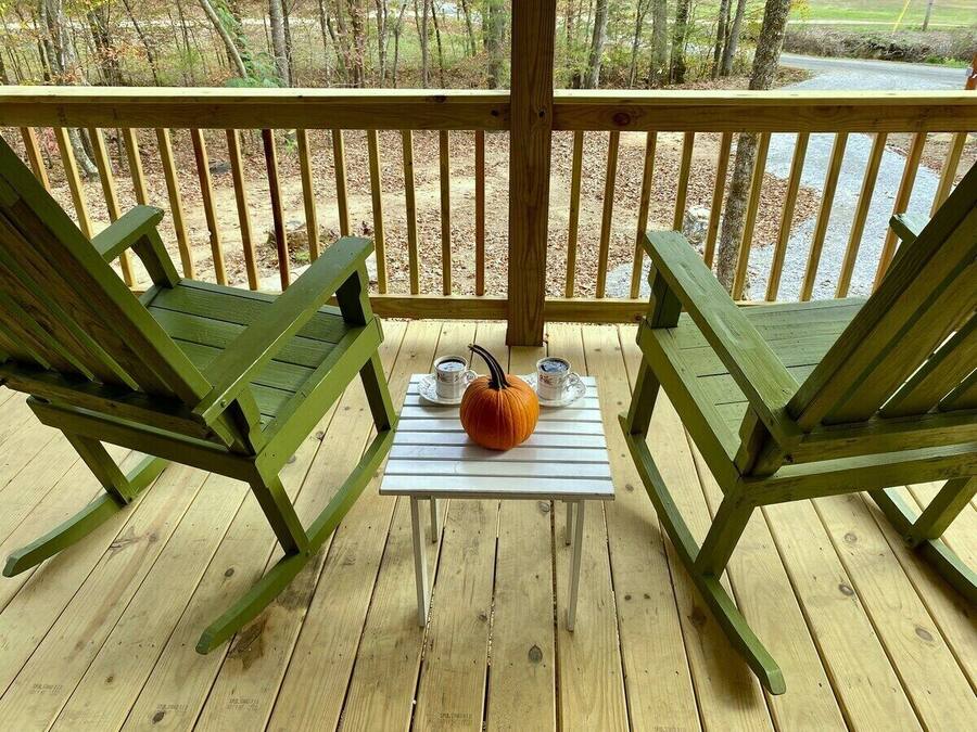 Une terrasse en bois avec deux fauteuils à bascule verts et une petite table sur laquelle se trouvent une citrouille et deux tasses.