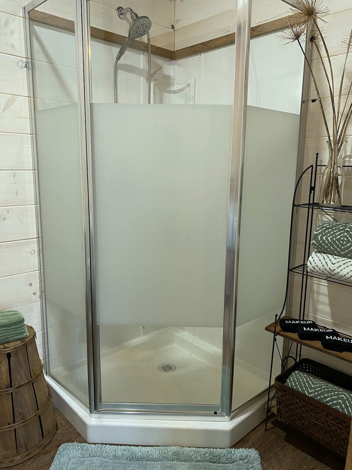 Doccia angolare con porta in vetro, uno sgabello di legno e uno scaffale con vari oggetti.
