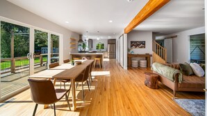 Amplia sala de estar con piso de madera, una mesa de comedor y un sofá. Se observa una cocina al fondo y vistas al exterior a través de amplias ventanas.