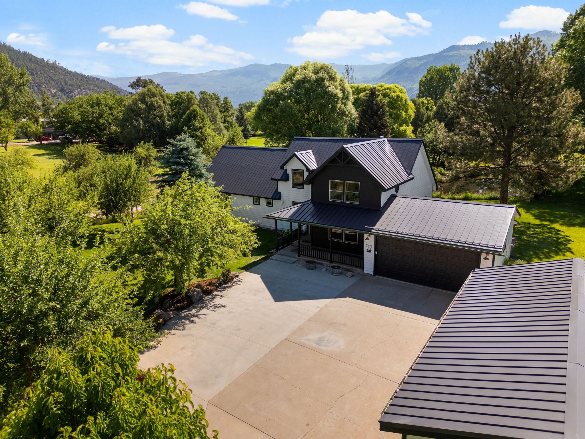Une maison à deux étages, dotée d’un porche abrité et d’un garage, entourée d’une végétation luxuriante et avec des montagnes en toile de fond.