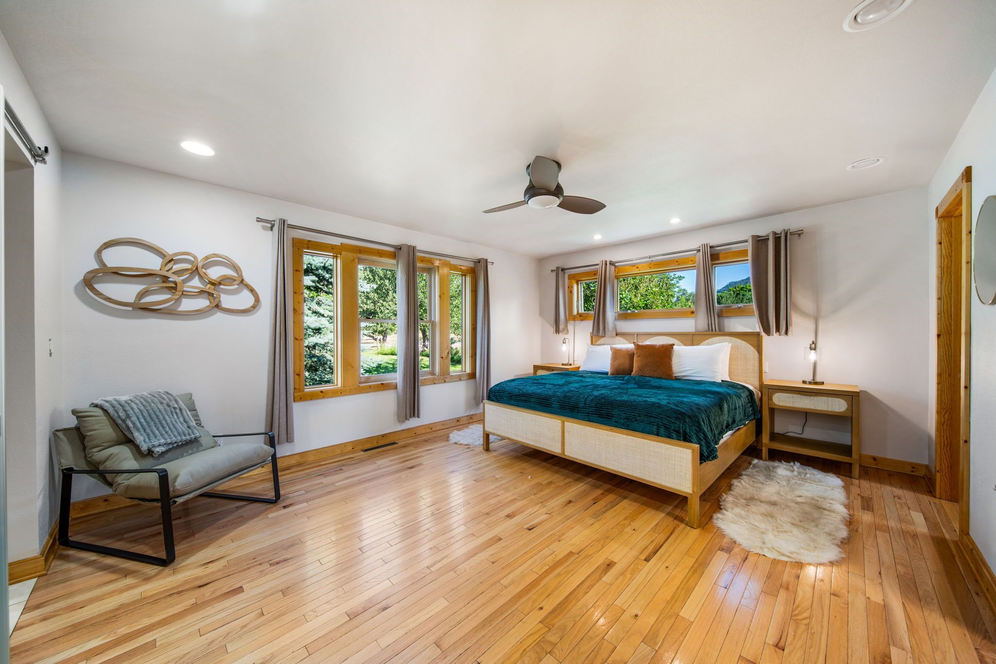 Une chambre avec un plancher en bois, un lit recouvert d’une couverture verte, une chaise, un ventilateur de plafond et de grandes fenêtres.
