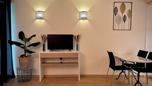 Un saló modern amb televisió de pantalla plana sobre un suport blanc, dues làmpades de paret, una taula de menjador amb cadires i una planta en test.