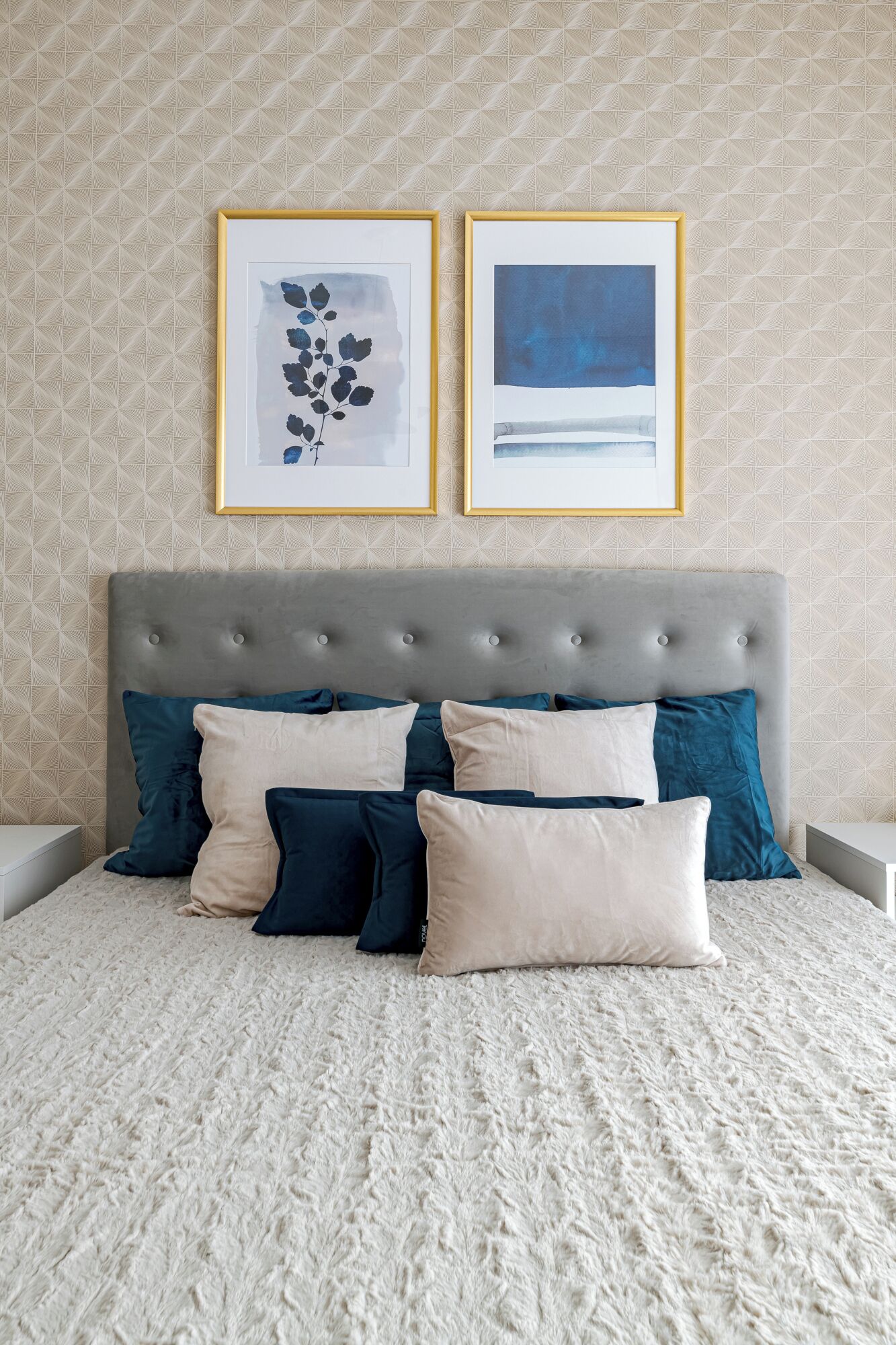 Un dormitorio con cabecera capitonada, papel tapiz estampado y dos obras de arte enmarcadas sobre la cama.