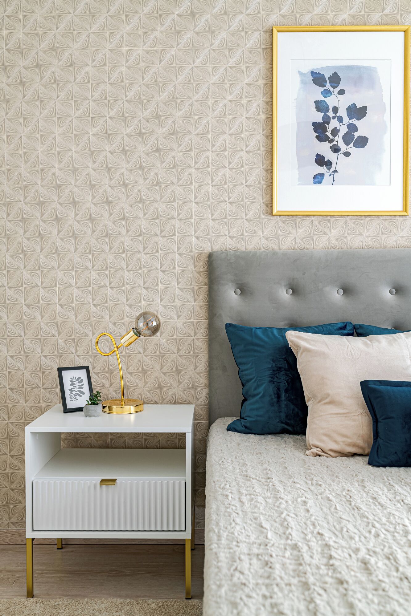 Een slaapkamer met behang met een patroon, een nachtkastje met een lamp, een ingelijst kunstwerk en een bed met meerdere kussens.