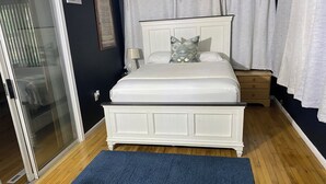 A bedroom with a white bed, a blue rug, a wooden nightstand, and a glass door leading to a balcony.