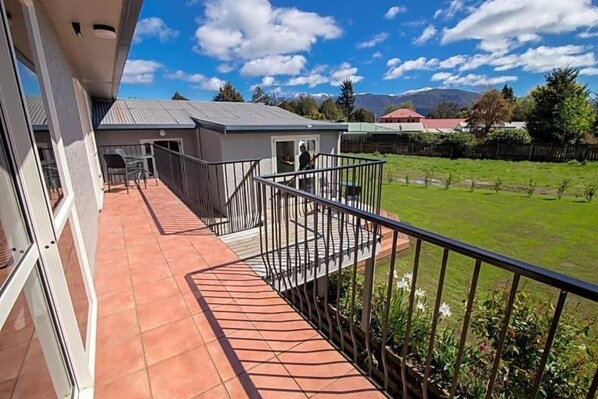 A balcony with a view of a green field and distant mountains, a small building with a red roof, and a clear blue sky. - Kea by the Lake (Te Anau)