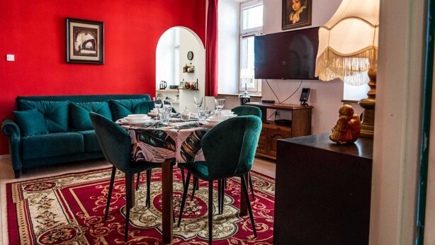 A dining area with a red wall, a teal sofa, a table set for four, and a TV in the background. - Górna Royal Red Apartment (Lublin)