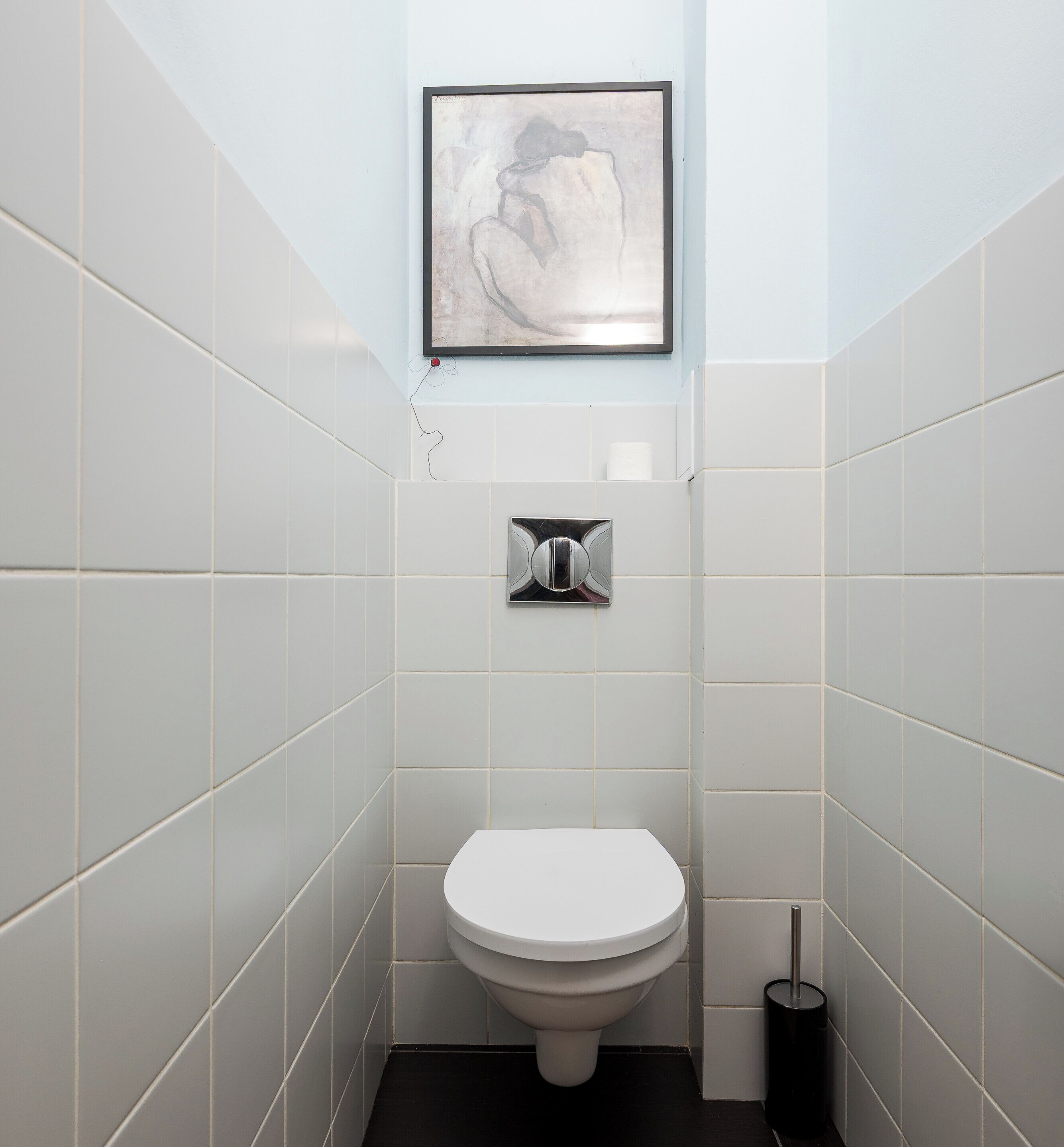 A small, modern bathroom with white tiles, a white toilet, a framed abstract artwork on the wall, and a trash bin.
