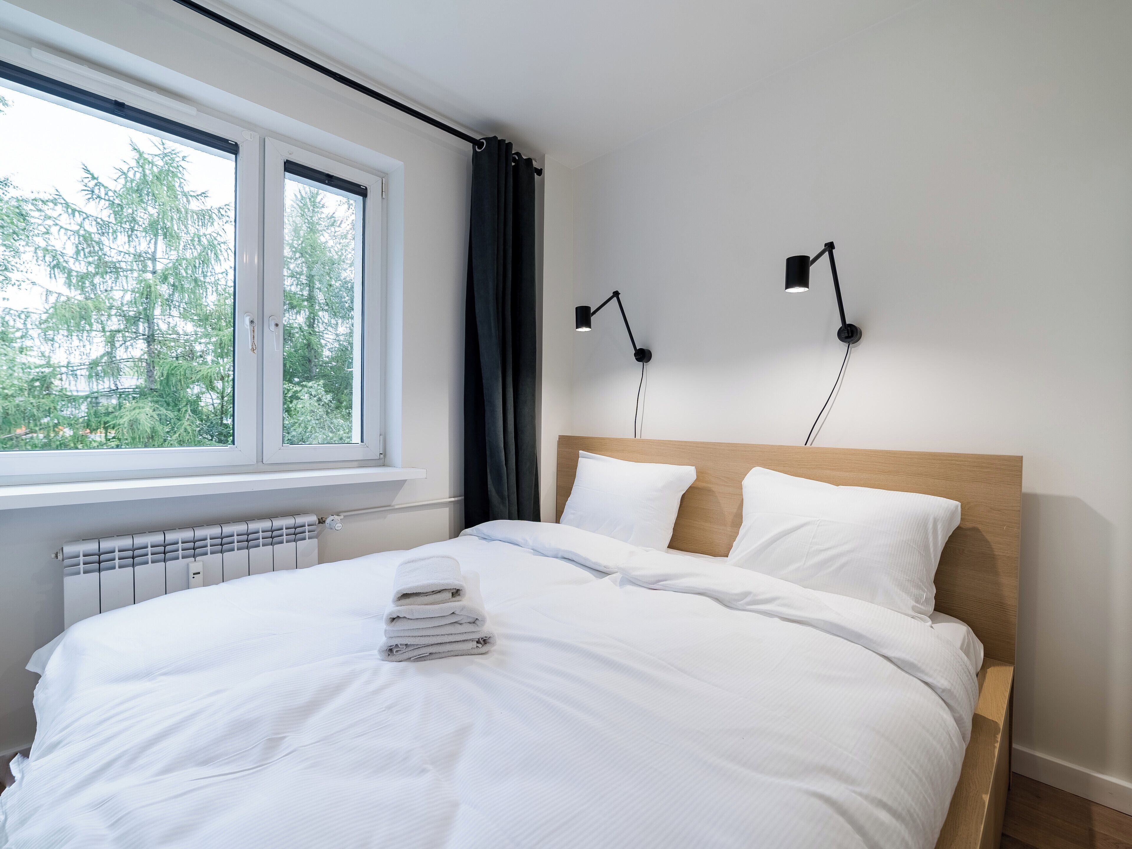 A neatly made bed with white linens and a stack of towels, a window with a view of trees, and two wall-mounted reading lamps.