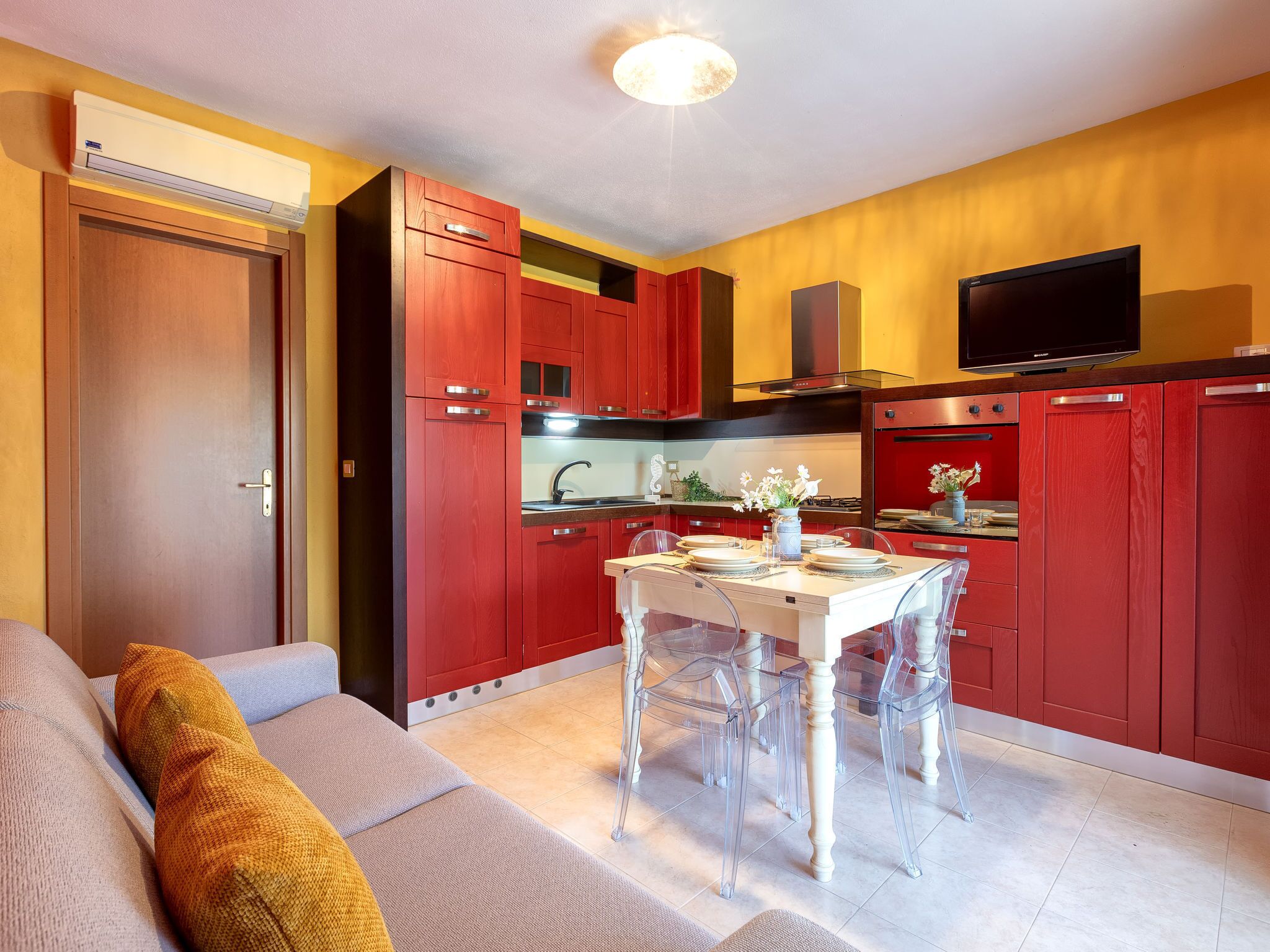 Une cuisine compacte avec des meubles rouges, une table à manger avec des chaises transparentes et un téléviseur à écran plat fixé au-dessus du comptoir.