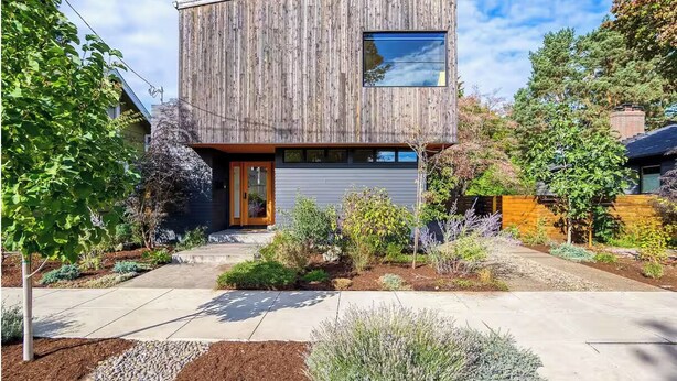 A modern two-story house with a wooden exterior, a front door, and a small garden with various plants and shrubs.