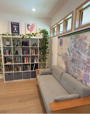 Ein gemütliches Wohnzimmer mit grauem Sofa, einer Bücherwand, einer Karte an der Wand und einem Fenster mit Holzrahmen.