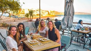 A group of people dining outdoors at a seaside restaurant with tables and chairs, overlooking the sea and buildings in the background. - Best room with WiFi in taghazout (agadir)