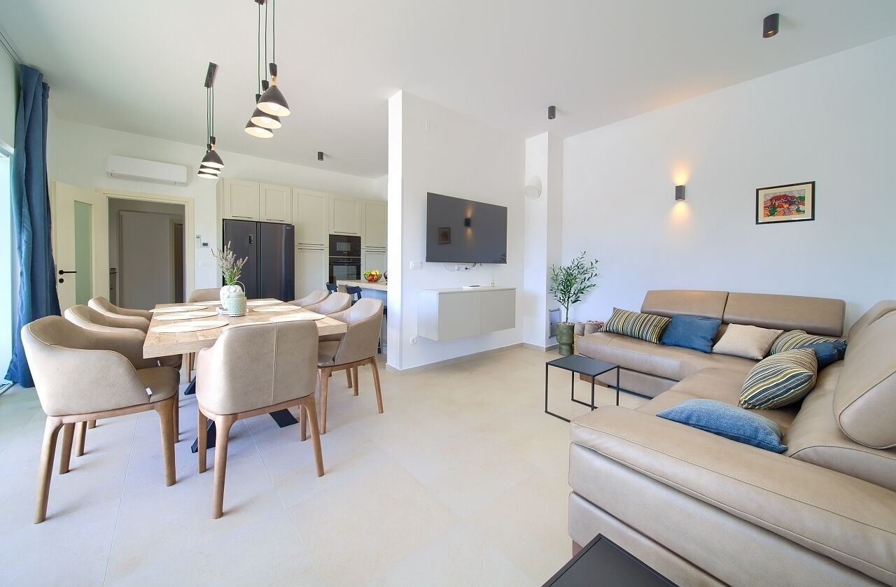 Ein modernes Wohnzimmer mit einer Couch, einem Esstisch und Stühlen. Ein Fernseher ist an der Wand befestigt, und im Hintergrund ist eine Küchenzeile erkennbar.