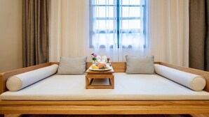 Ein Zimmer mit einem hölzernen Bett, beigen Kissen, einem kleinen Holztisch mit einer Essschale und einem Fenster mit Vorhängen.