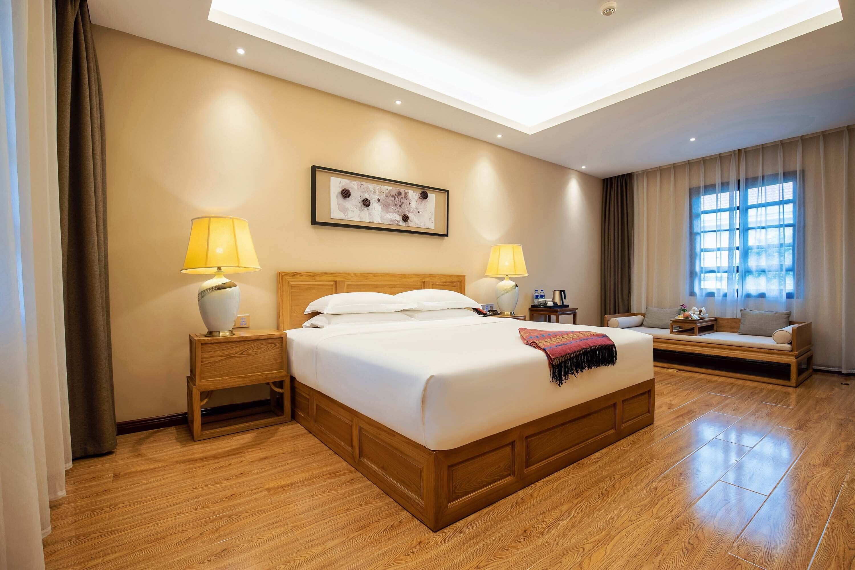 A hotel room with a large bed, two bedside lamps, a wooden headboard, a window with curtains, and a small seating area with a sofa and a table.