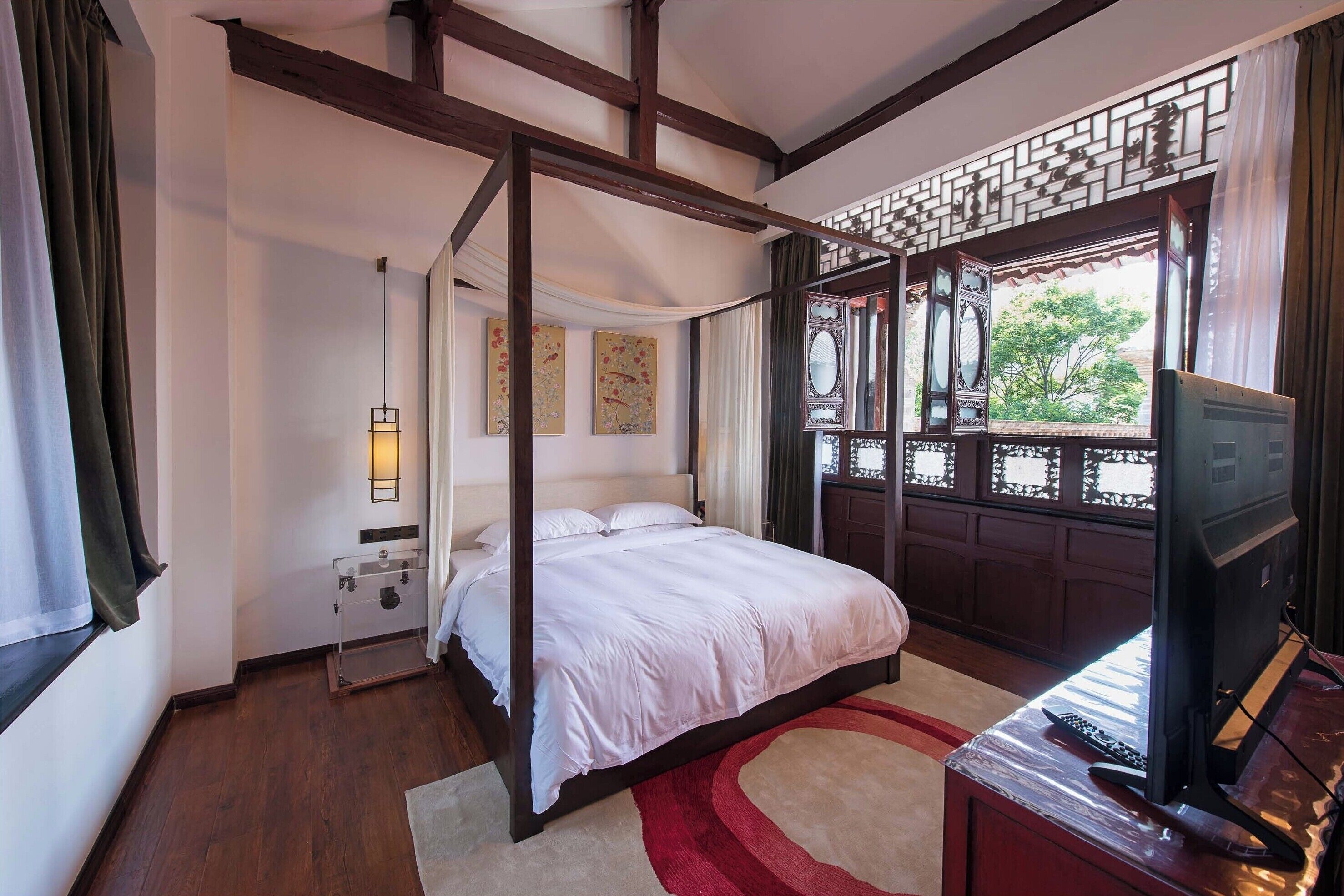 A bedroom with a four-poster bed, a television, and a window with a view of greenery.
