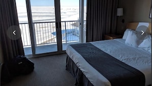A hotel room with a bed, a suitcase, a nightstand, a lamp, and a view of the beach through a sliding door. - Top floor Lakeview Hotel 1 Bd available July 31 - Aug 7 Icelandic festival week (Gimli)