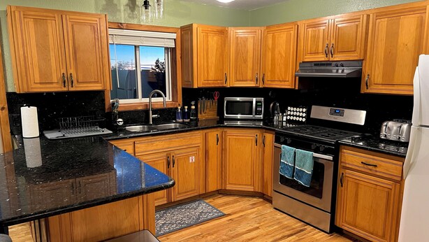 Une cuisine avec des meubles de rangement en bois, un plan de travail noir, un évier en acier inoxydable, un four à micro-ondes, une cuisinière et un réfrigérateur.