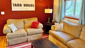 Un salon avec deux canapés, un ventilateur de plafond, une enseigne indiquant « TAOS HOUSE, altitude 7 123 pieds », et une fenêtre avec des rideaux.