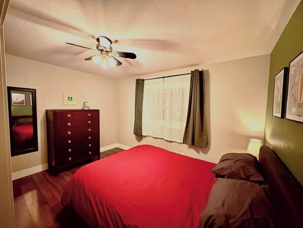 Una camera da letto con un letto, una cassettiera, una ventola a soffitto, una finestra con tende e quadri appesi al muro.