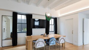 Un comedor moderno con una mesa de madera y sillas tapizadas, un televisor de pantalla plana en la pared y un ventanal con cortinas negras.