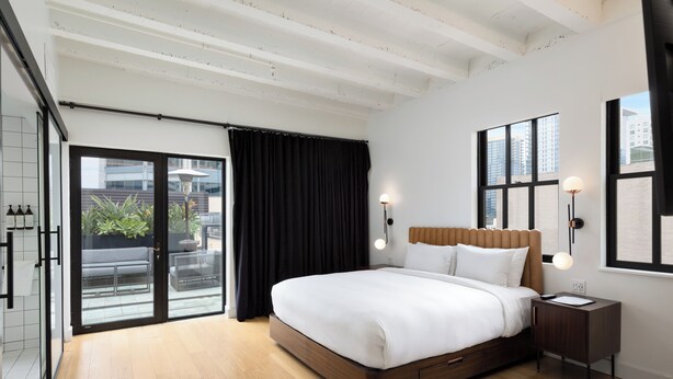Un dormitorio moderno con una cama grande, suelo de madera, puerta de vidrio corrediza que da a un espacio exterior y vistas a la ciudad a través de las ventanas.
