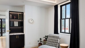 Un salón moderno con un sillón a cuadros blanco y negro, una mesita redonda de madera, una estantería con libros y objetos decorativos, una planta en maceta y un reloj de pared.