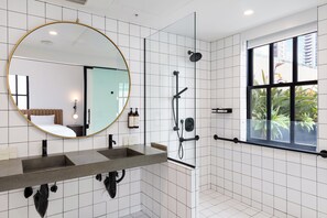 Un baño moderno con un espejo grande, dos lavamanos, una ducha y una ventana con vistas a zonas verdes y edificios.