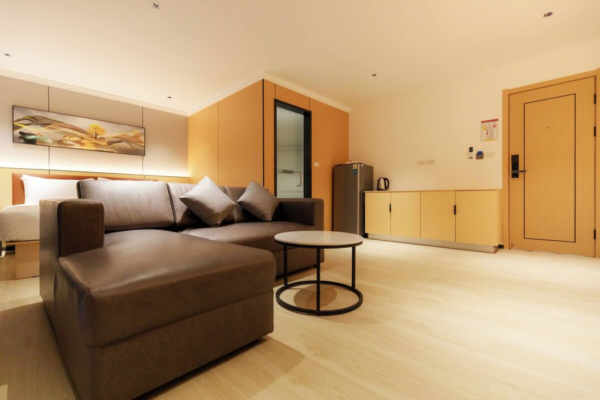 A modern living room with a brown sectional sofa, a small round table, and a kitchen area with a refrigerator and cabinets.