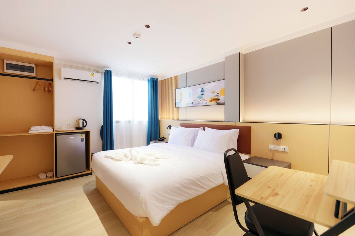 A hotel room with a bed, a desk, a chair, a small refrigerator, a bedside table, a wall-mounted air conditioner, and a window with curtains.