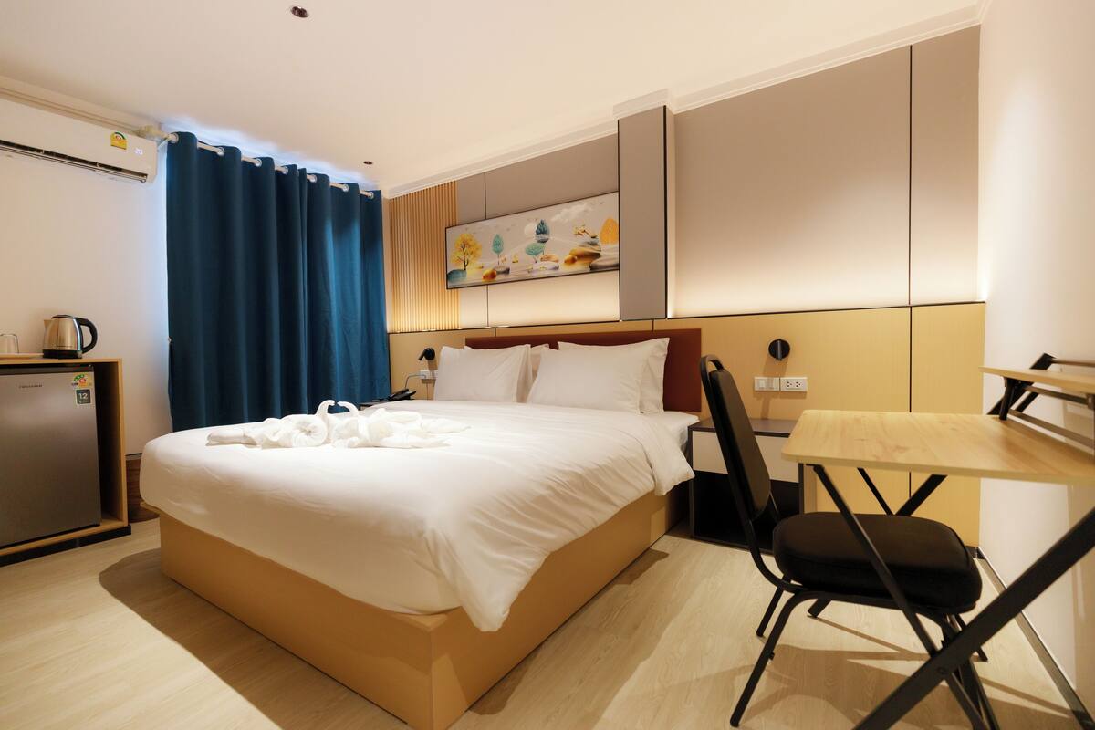 A hotel room with a bed, a desk, a chair, a painting on the wall, and a small kitchenette.
