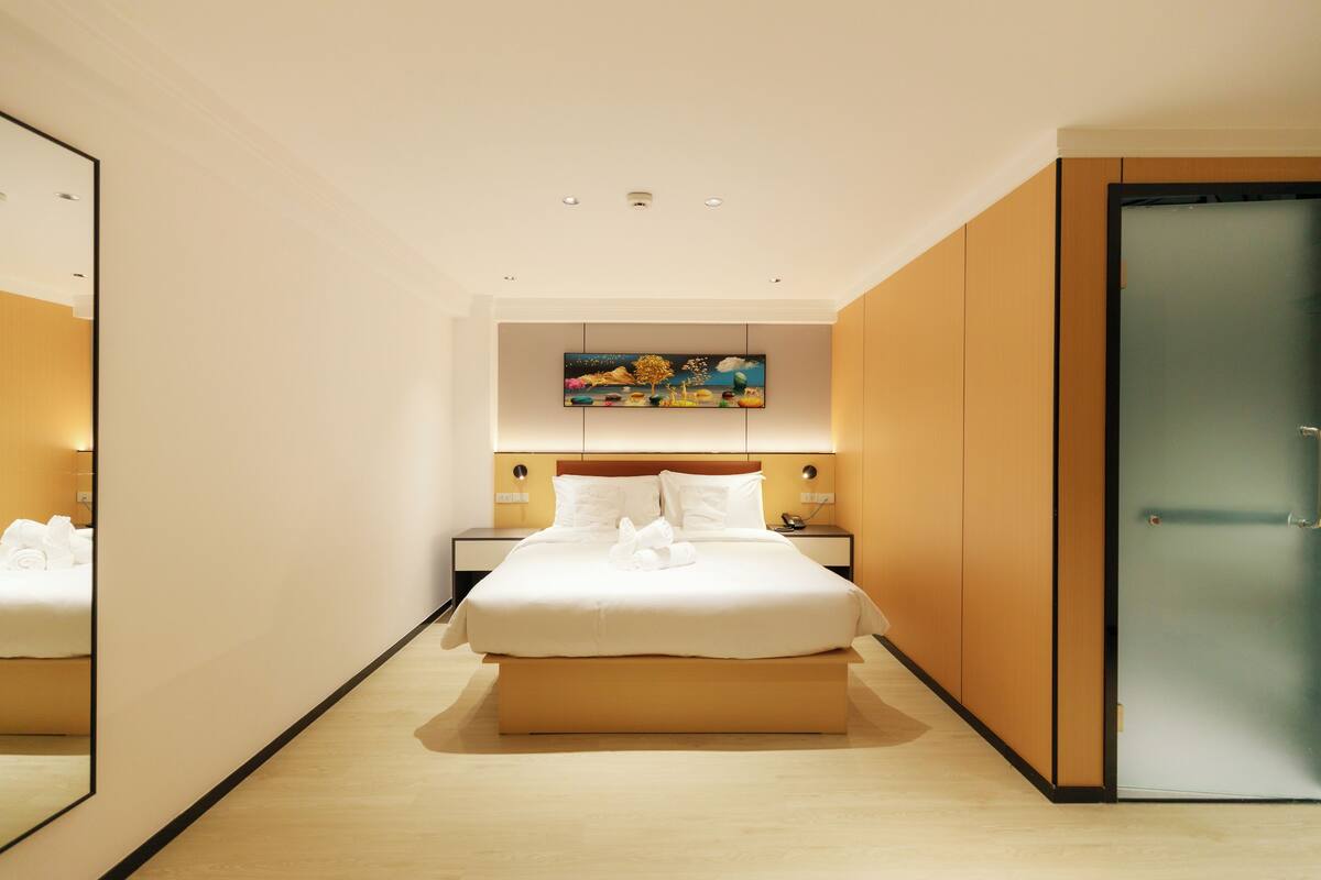 A modern hotel room with a large bed, a painting above the headboard, and a glass door on the right.