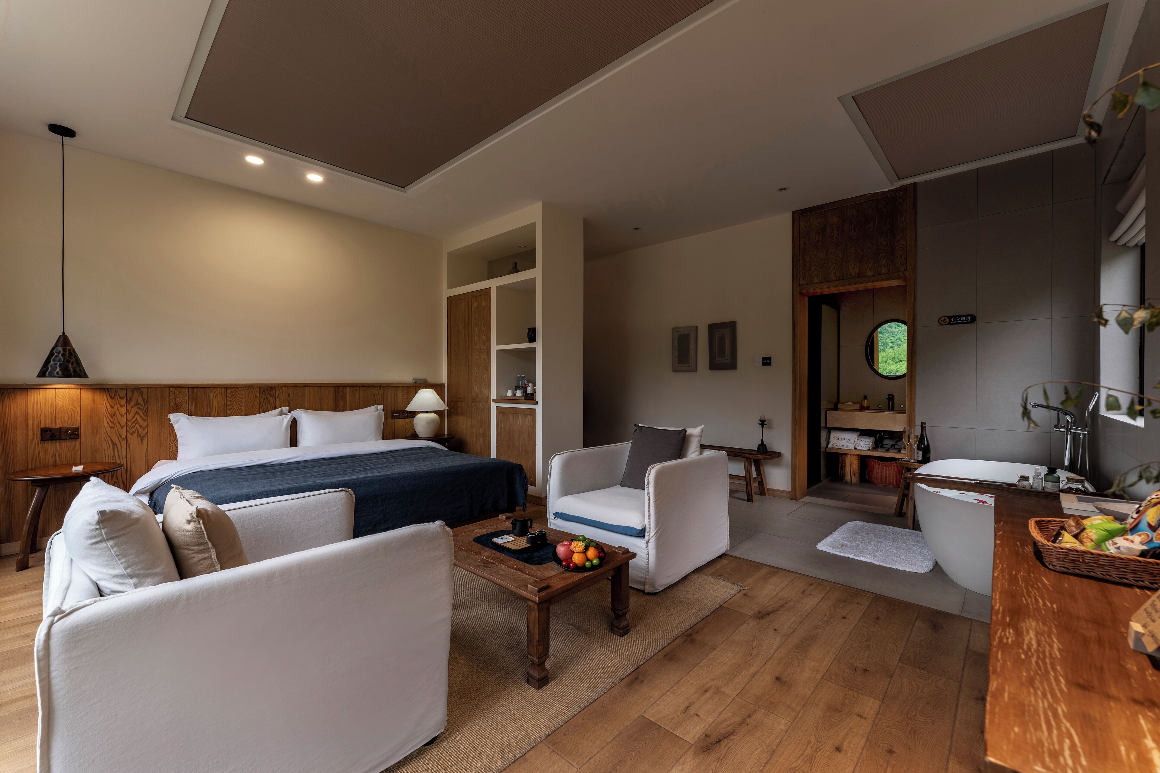 A modern hotel room with a bed, sofa, armchair, and a small table with fruit. There is a bathroom with a bathtub and a sink.