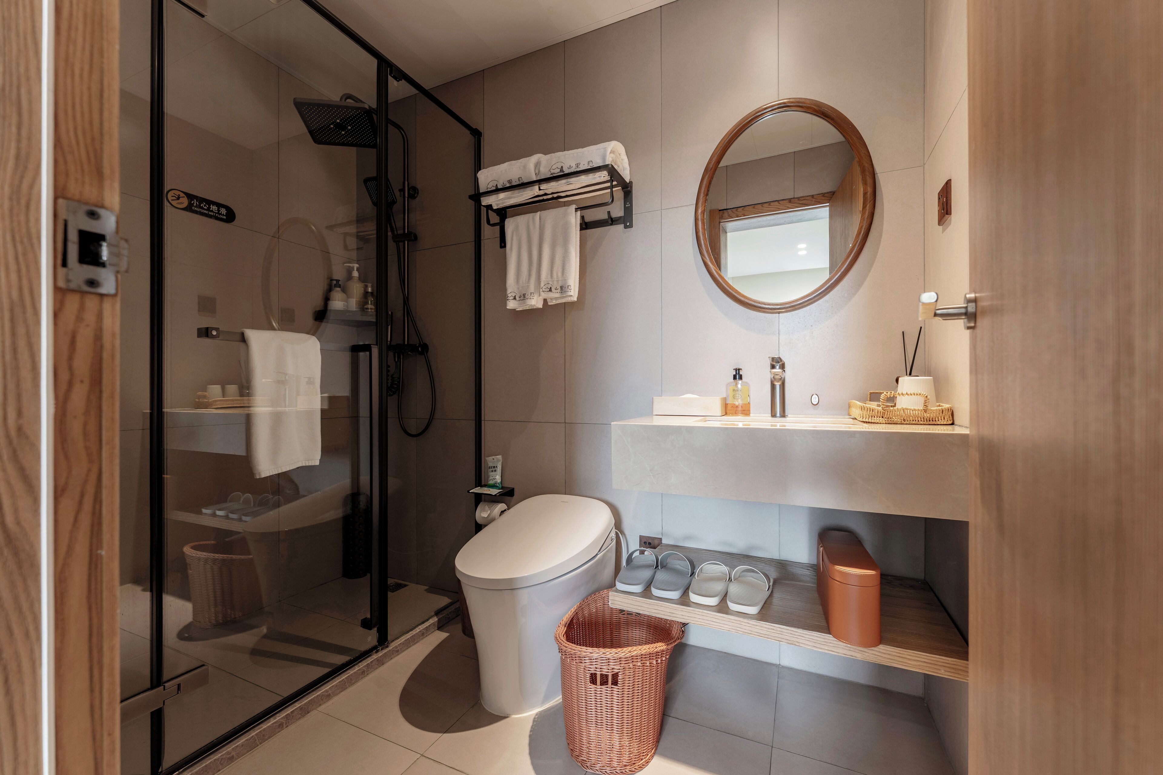 A modern bathroom with a glass shower enclosure, a toilet, a sink with a mirror, and a storage shelf with toiletries.