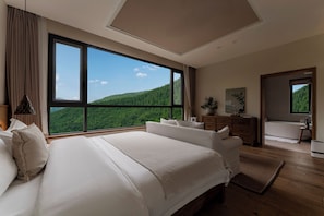 A bedroom with a large bed, a dresser, a vanity, and a view of a forested mountain.