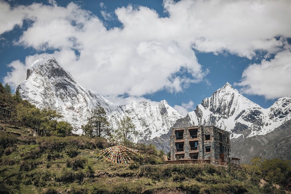 A stone building with multiple windows and a wooden deck, surrounded by snow-covered mountains and a clear sky.